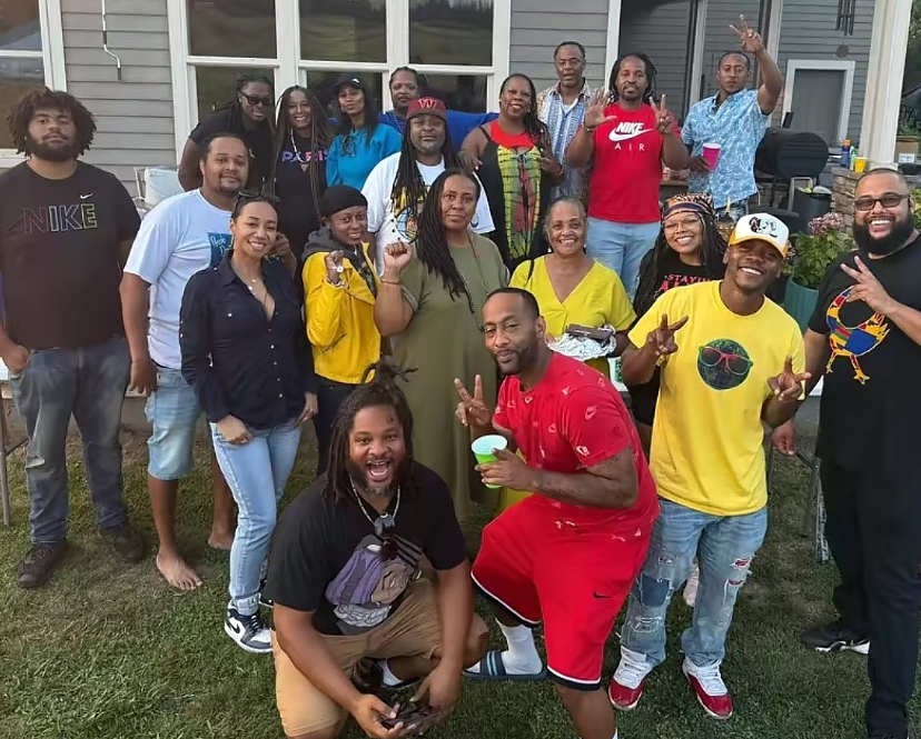 Group of people smiling outdoors, near a house. Some are holding drinks or food.