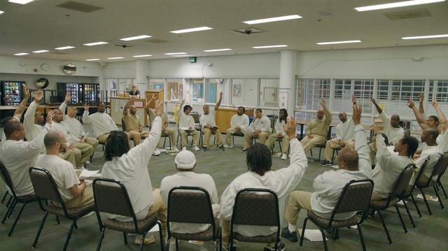 People in a circle raising hands, in a room with windows and artwork, likely a group discussion.