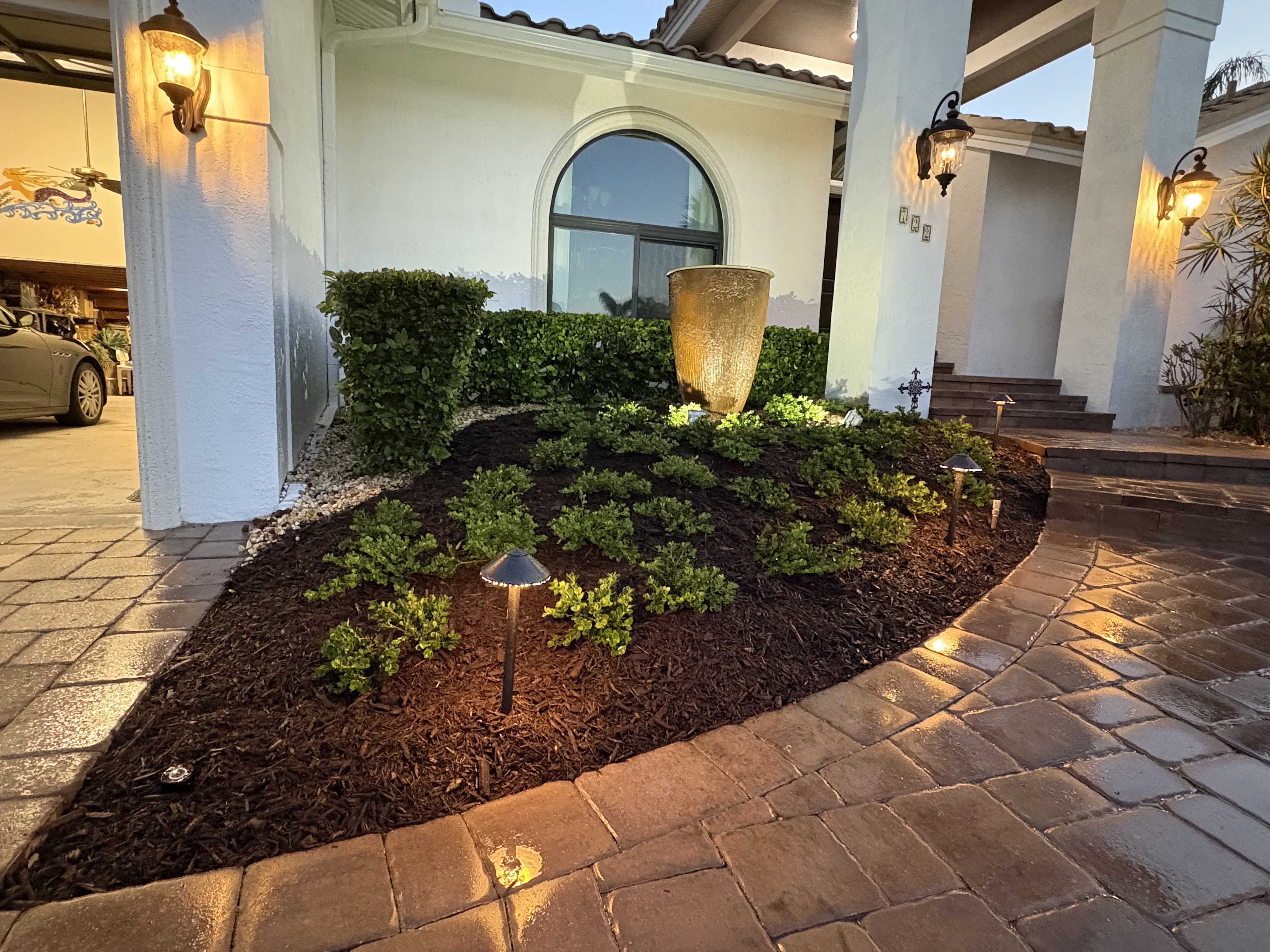 Front entrance of a house with a brick walkway, landscaped garden with lighting, and a water fountain. The stucco exterior has a curved arch.