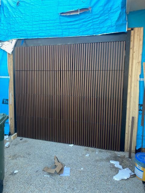 Closed brown metal garage door under a blue tarp-covered structure, with debris on the ground.