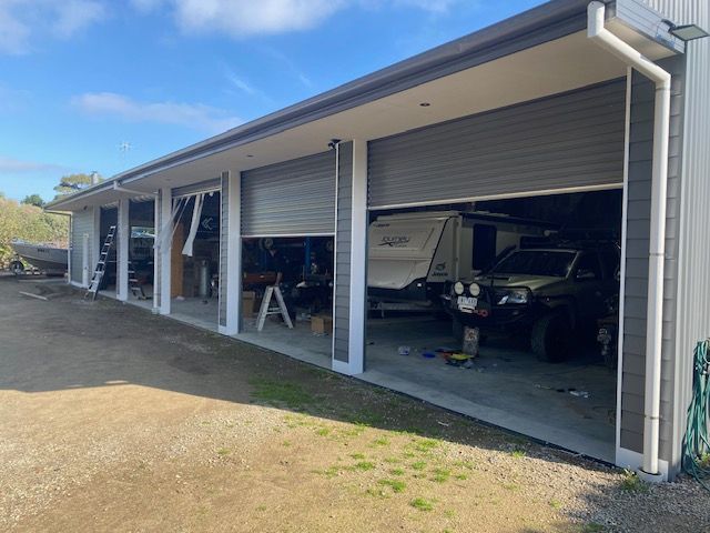 Open garage bays with a dark SUV and tools inside, viewed from a gravel driveway on a sunny day