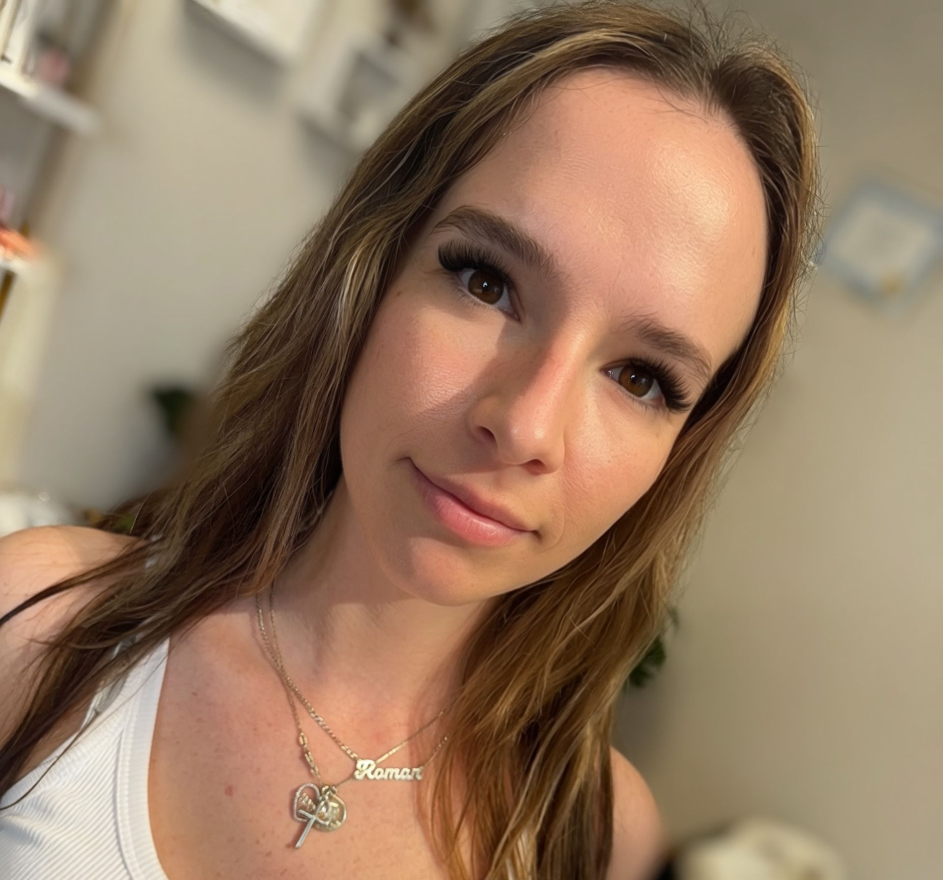 Woman with long lashes and light brown hair wearing a white tank top and gold necklace, looking at the camera.