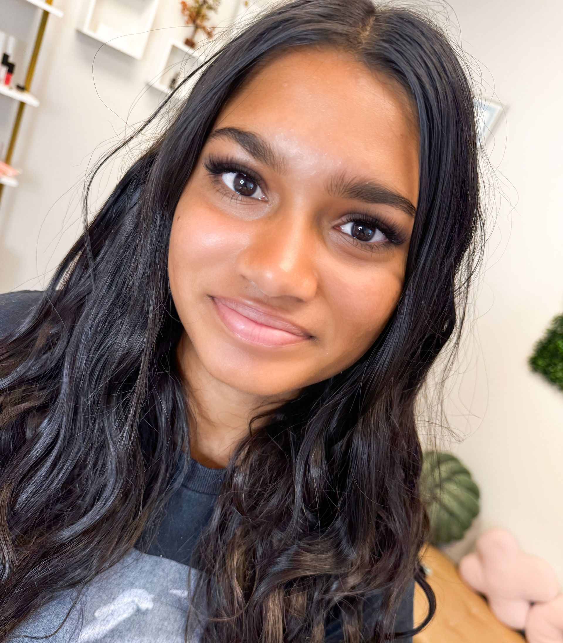 Woman with long dark wavy hair smiles, wearing dark shirt; indoor setting, eyelash extensions visible.
