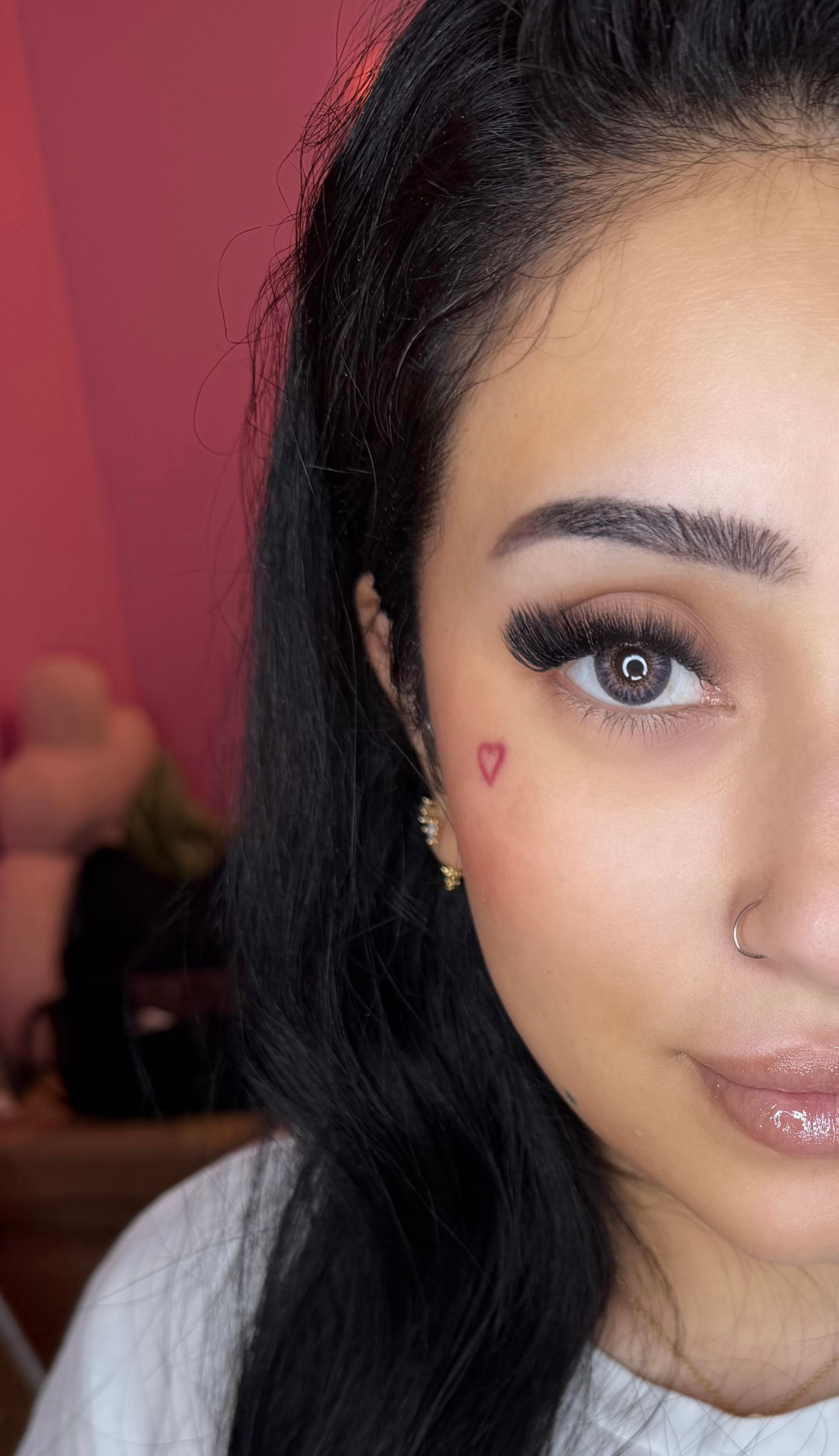 Woman with dark hair, heart cheek tattoo, and long lashes, posing for a selfie. Pink background.