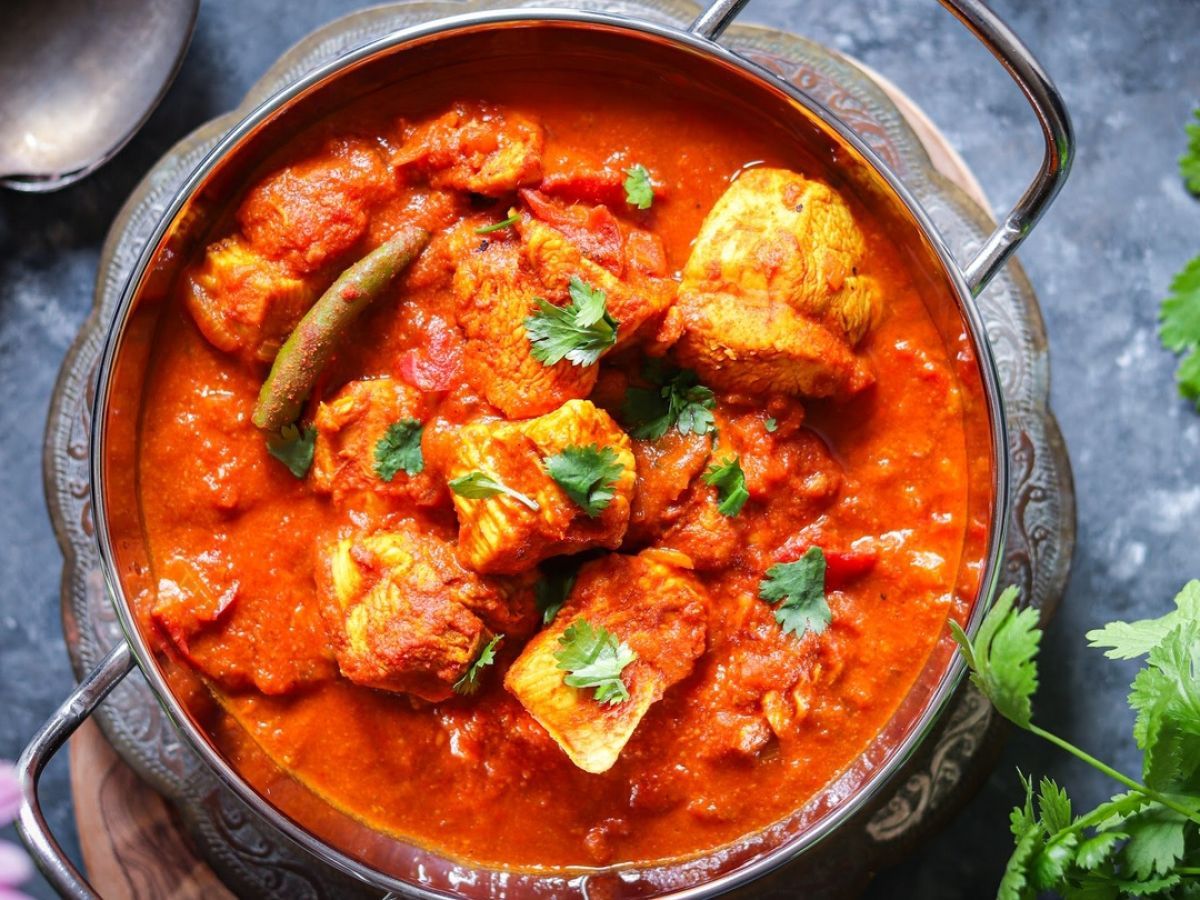 Un bol métallique rempli d'un curry de poulet rouge vif, garni de coriandre fraîche sur un fond texturé foncé.