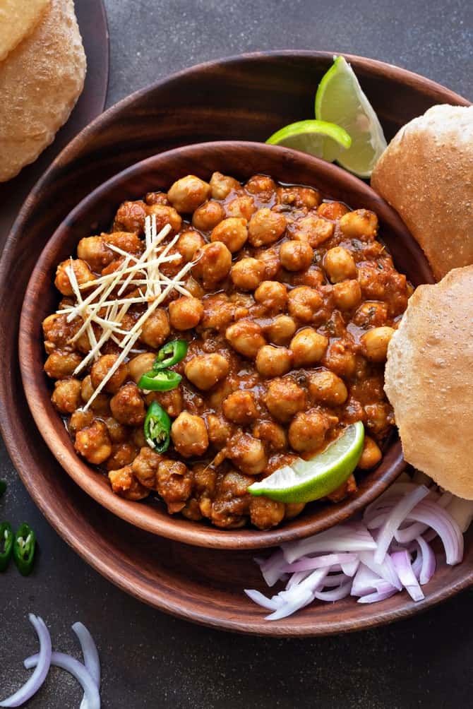 Un bol de chana masala garni de gingembre et de piments verts, servi avec du pain frit, des quartiers de citron vert et des oignons émincés.