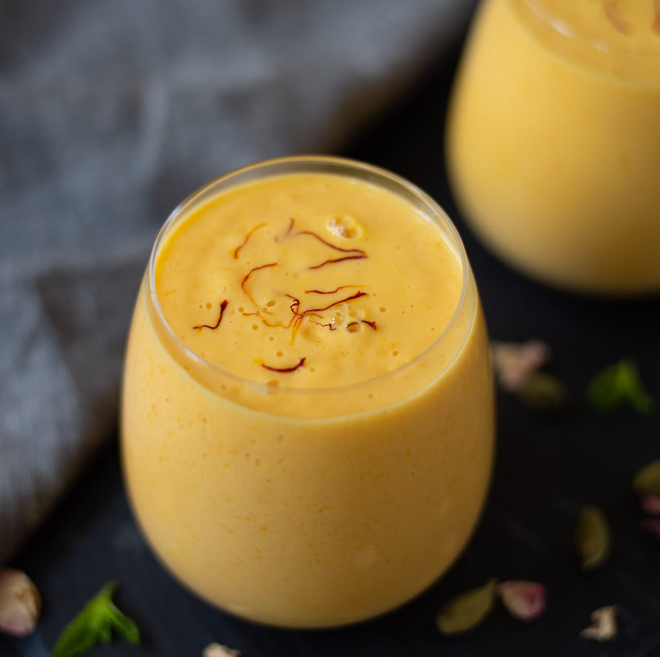 Deux verres de lassi crémeux à la mangue, garnis de filaments de safran, posés sur une surface sombre parsemée de pistaches et de pétales.