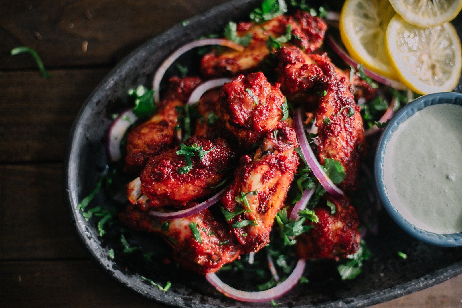 Un plat sombre d'ailes de poulet rouges glacées, garnies d'oignons rouges émincés, d'herbes et de citron, accompagnées d'une sauce crémeuse.