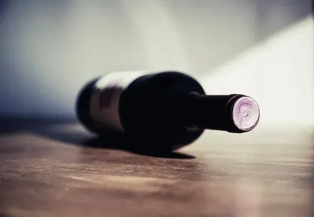 Une bouteille de vin en verre foncé, fermée par un bouchon de liège recouvert d'une capsule en aluminium, repose horizontalement sur une surface en bois dans une lumière tamisée.