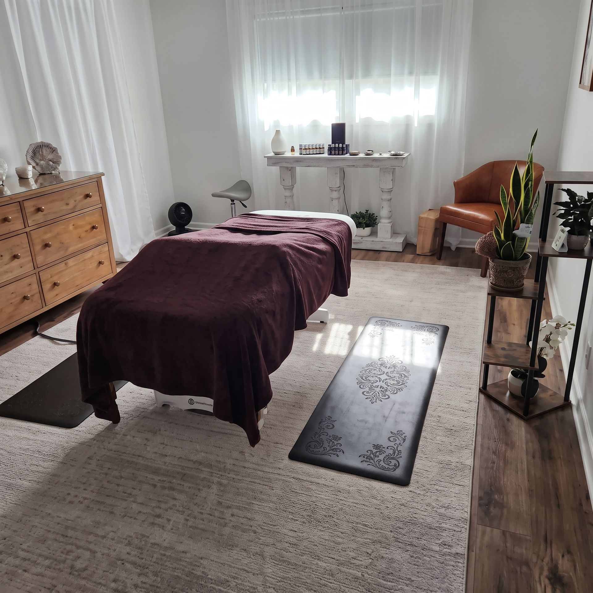 Bright massage room with a treatment table, rug, dresser, chair, and plants by a window