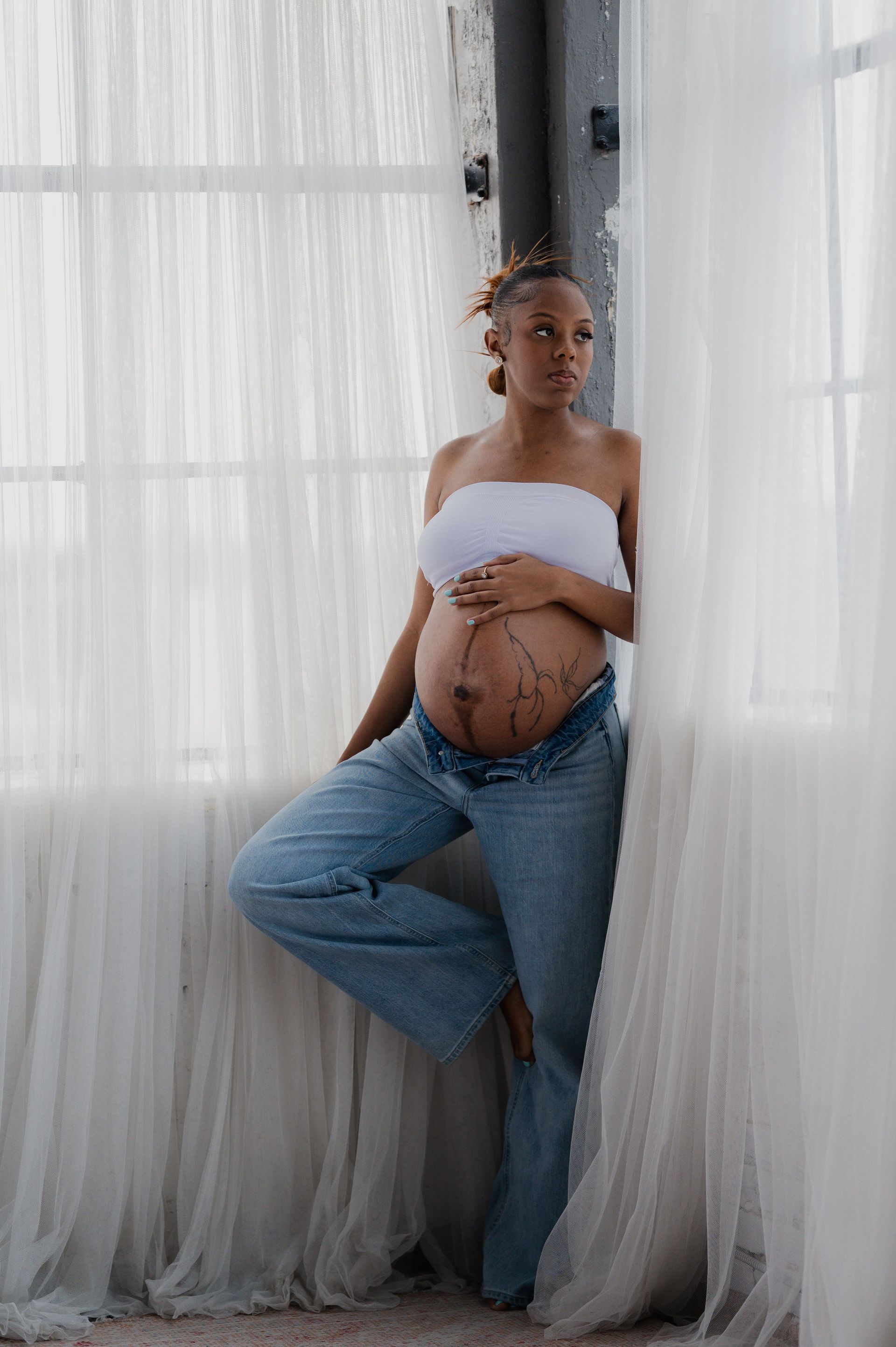 Pregnant woman in a white top and jeans standing by sheer curtains in soft natural light