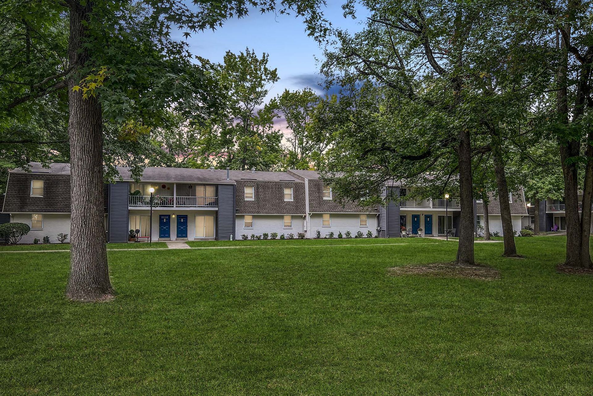 Exterior view of a two-story apartment building with blue doors and a grassy, tree-filled courtyard.