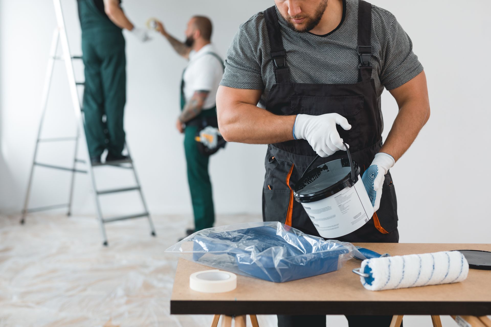 Painter pouring paint into a tray while coworkers paint walls in the background. Painter pouring paint into a tray while coworkers paint walls in the background.