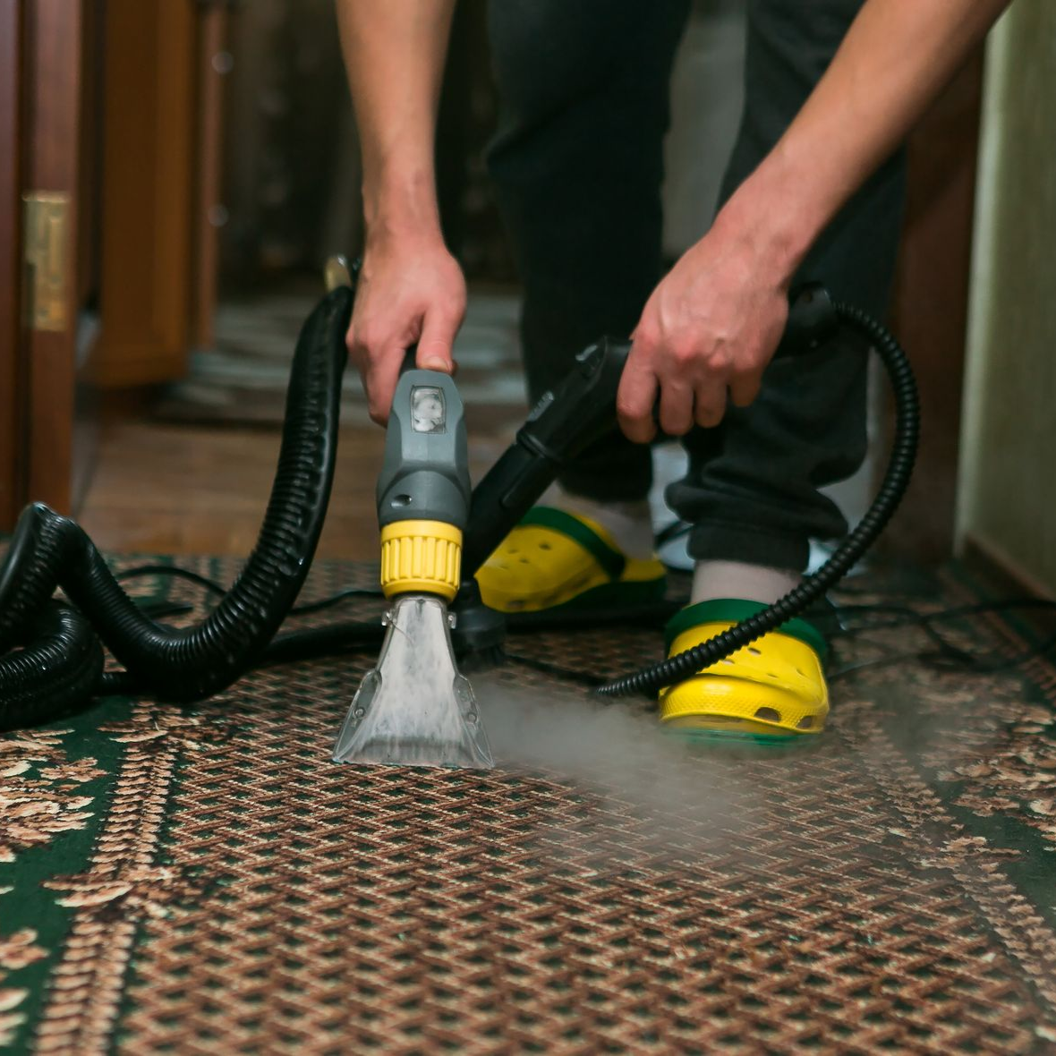 Person vacuums a white rug with a geometric pattern in a living room. A yellow vacuum cleaner is nearby.
