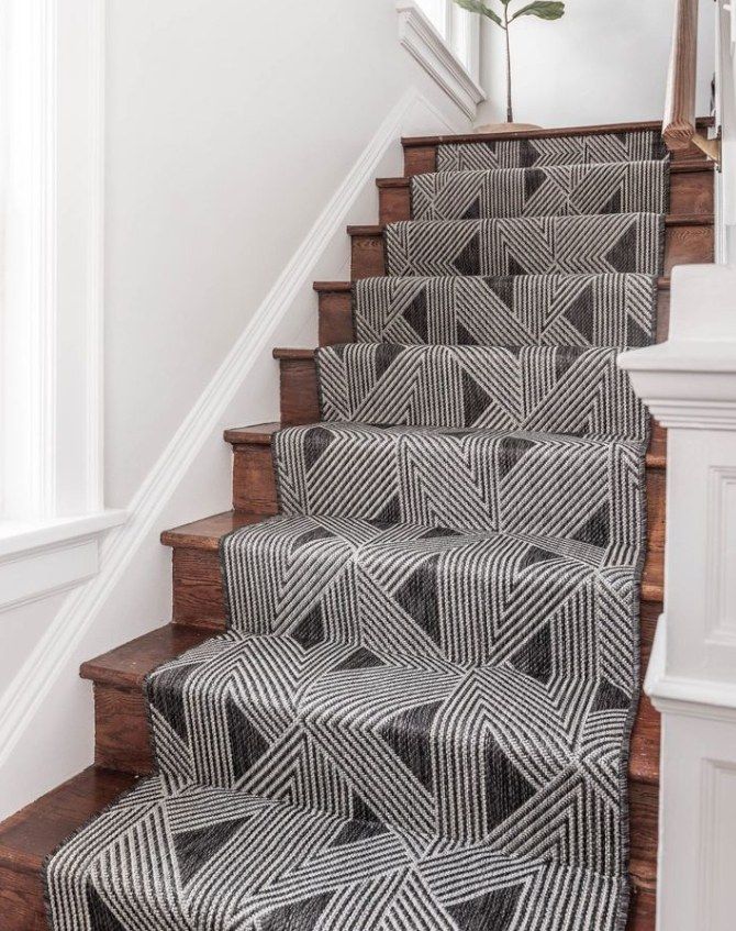 Staircase with patterned gray and black carpet runner. Brown wooden steps and white walls.