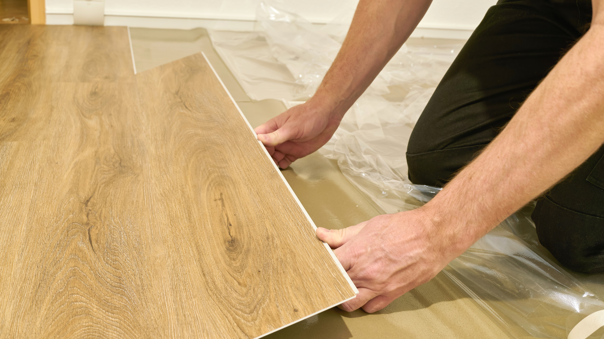 Person installing wood-look flooring, kneeling and aligning planks on plastic sheeting.