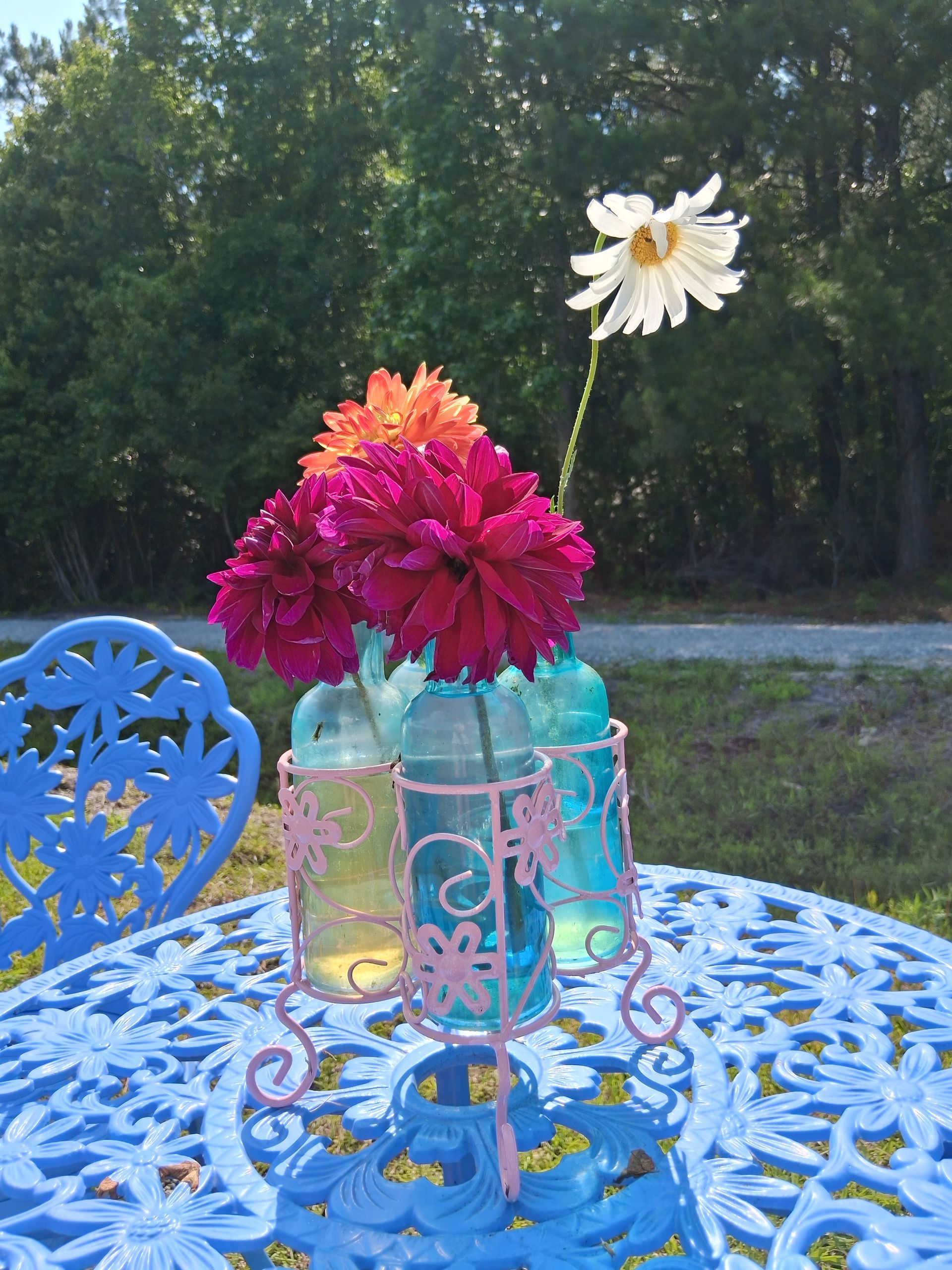 Bright dahlias and a white daisy in blue glass bottles on a pink wire stand atop an ornate blue metal table outdoors.