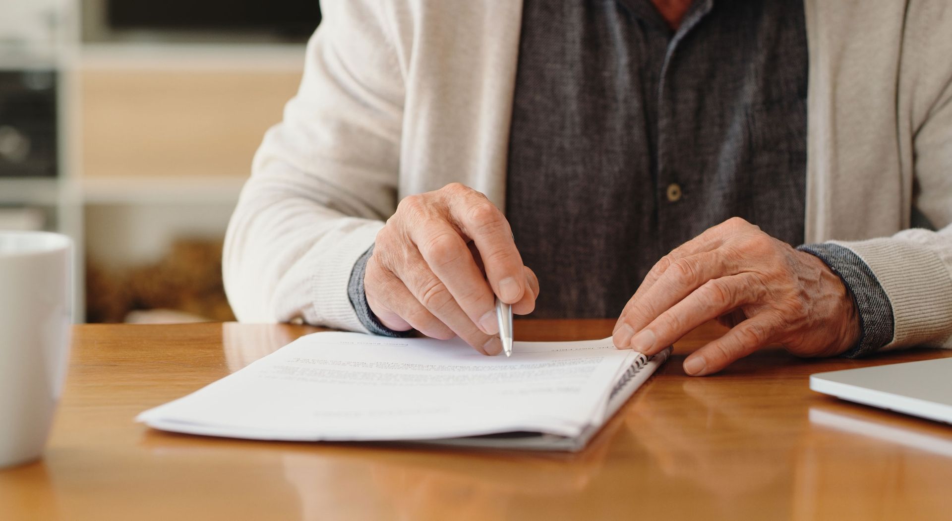 Person signing document at wooden table, with pen in hand. Beige sweater and gray shirt visible.