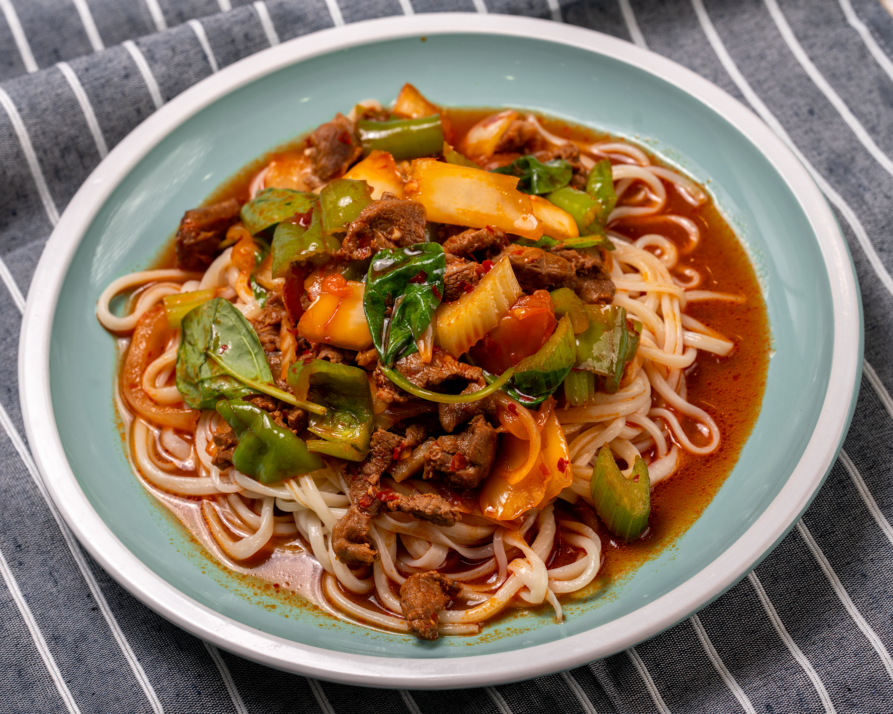 Noodles in a bowl topped with stir-fried vegetables and sauce on a striped cloth