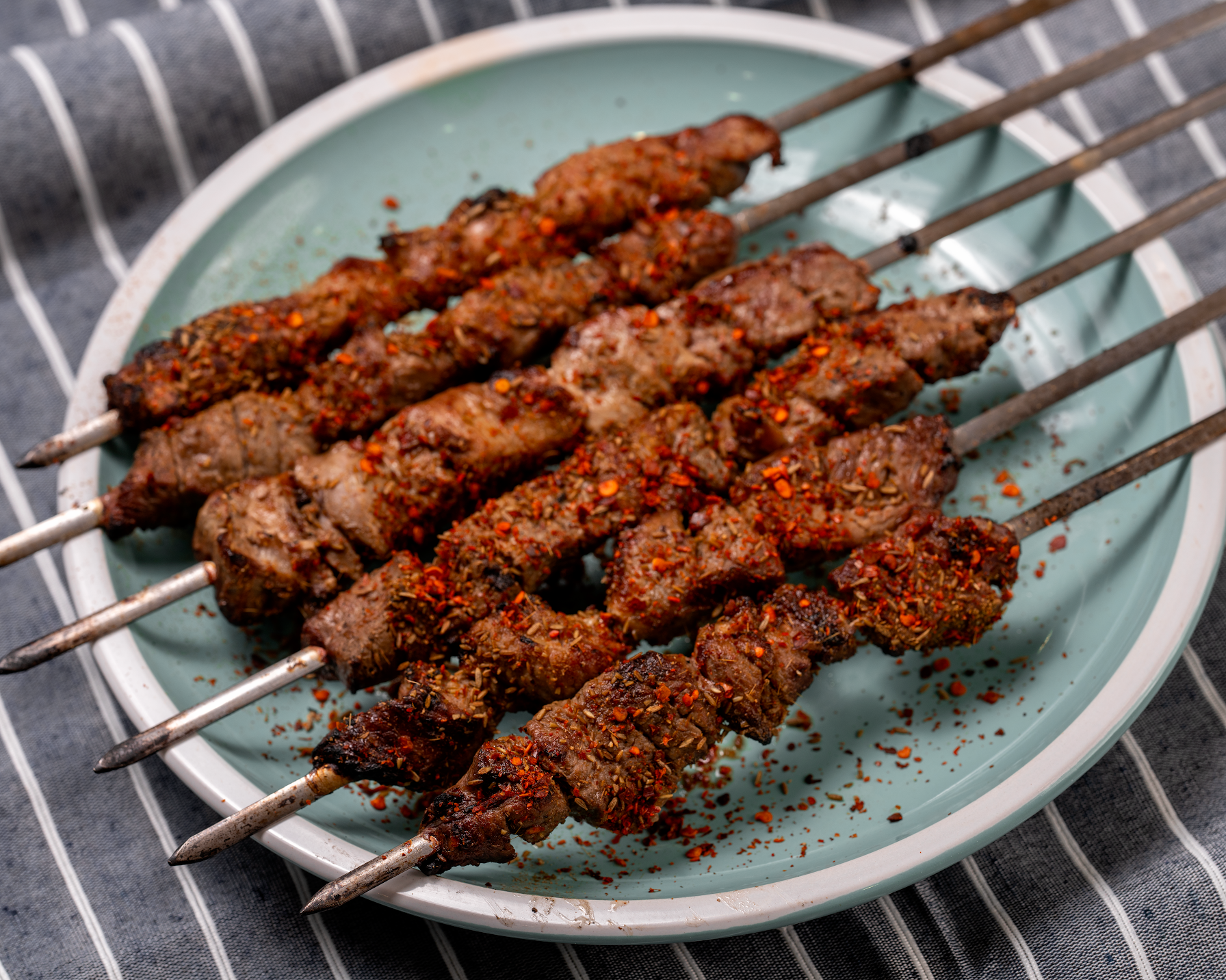 Grilled spiced meat skewers on a light blue plate with red seasoning on a striped cloth