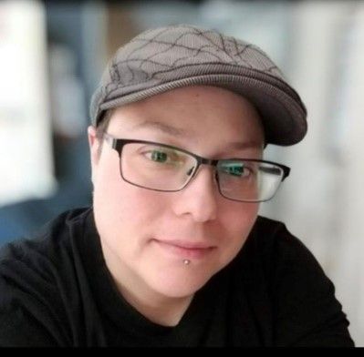 Person wearing glasses and a hat, looking at the camera. They have a lip piercing and are wearing a black shirt.