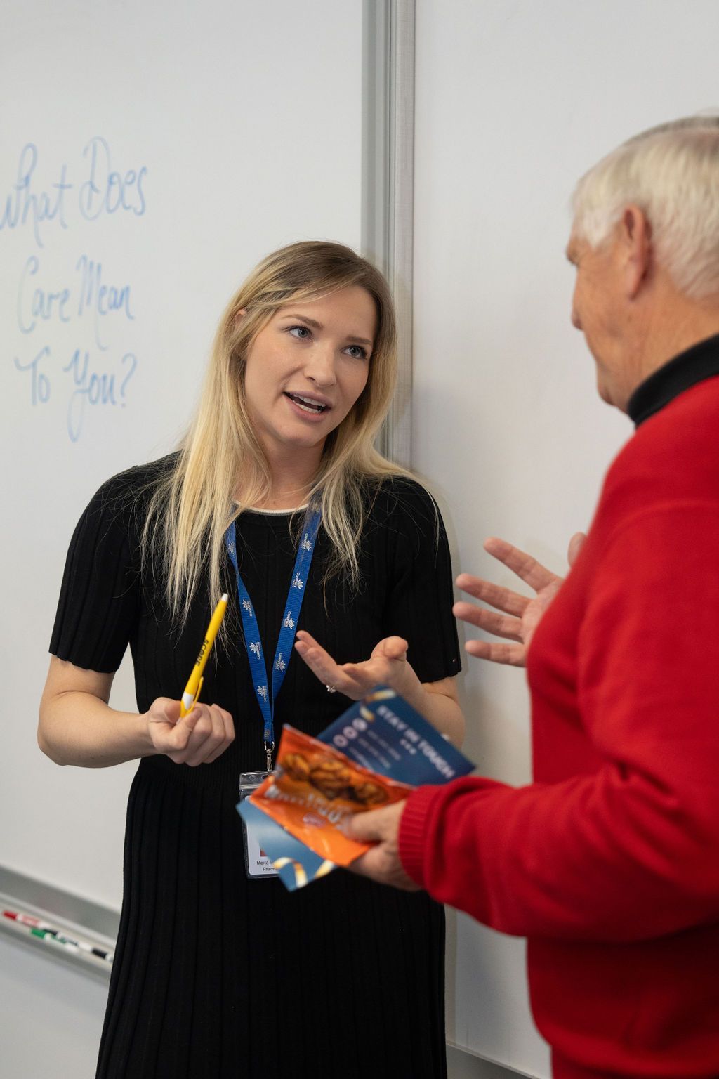 A woman in a black dress is talking to a man in a red sweater in front of a white board.
