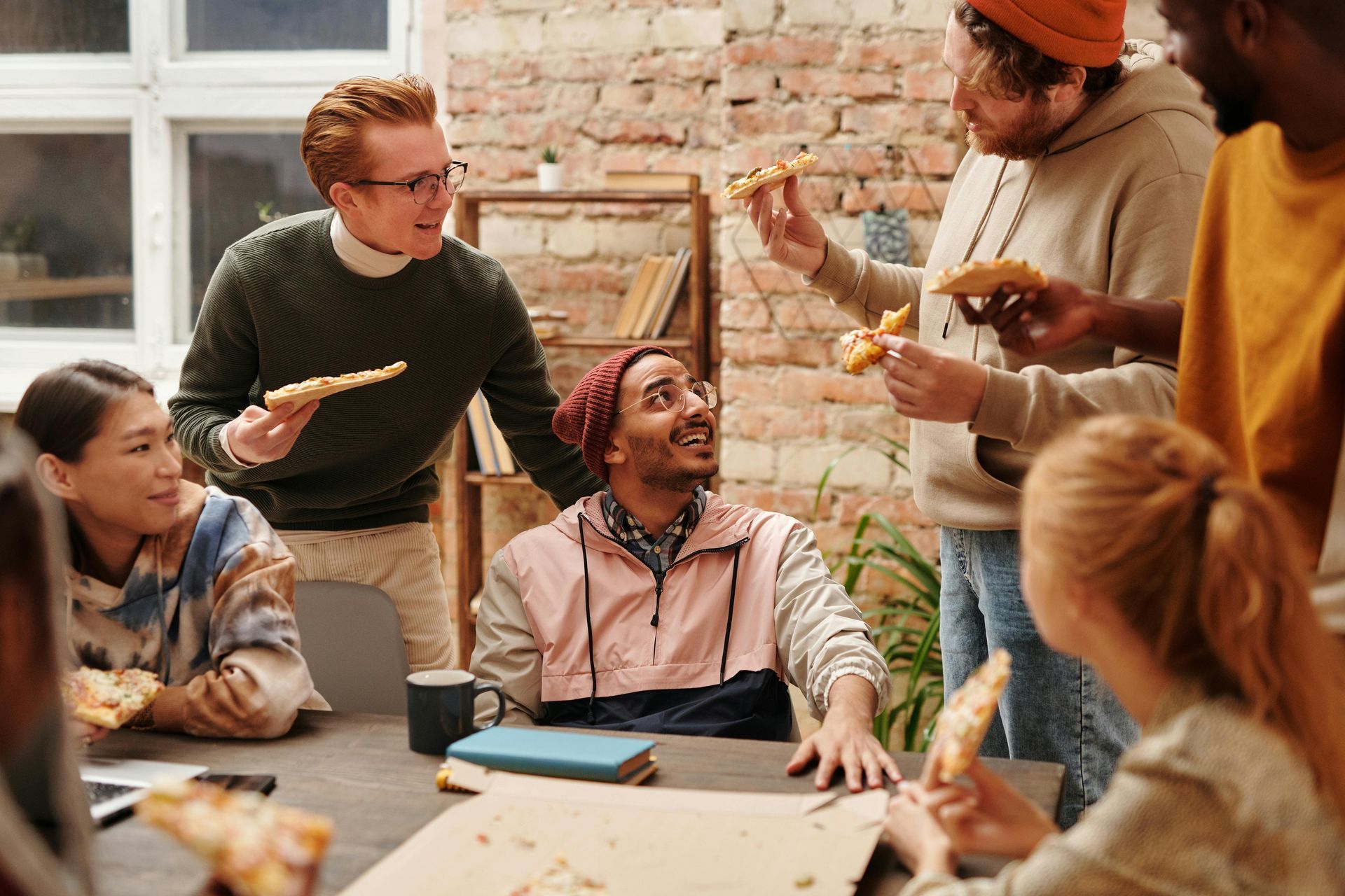 Group of people eating pizza around a table in a brick-walled room. They are talking and smiling.