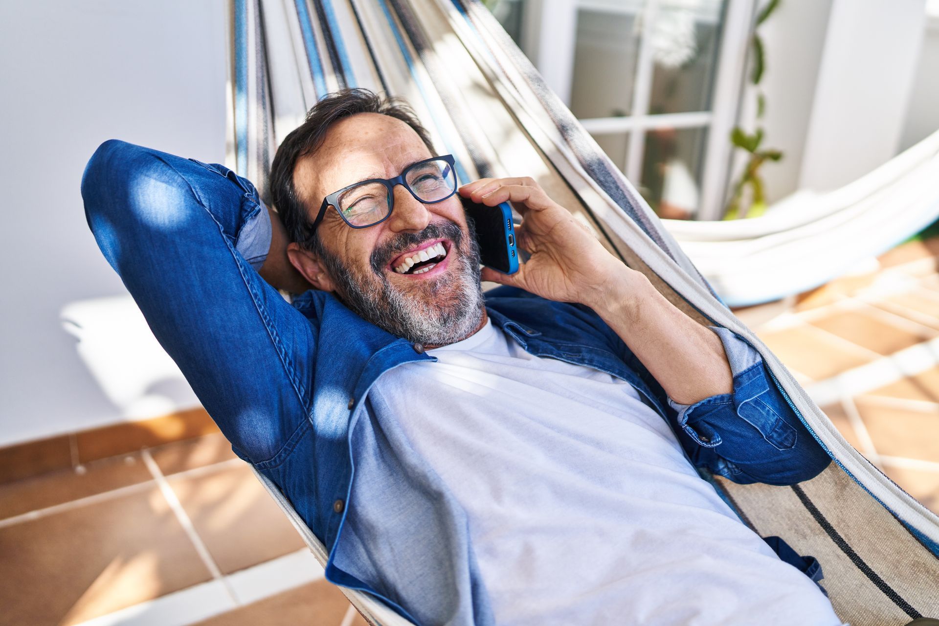 Smiling man in glasses reclining in a lounge chair, talking on a phone on a sunny patio