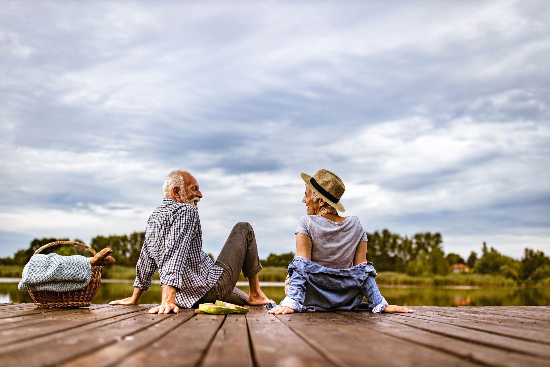 Two people sit on a wooden dock facing a cloudy lake, with a basket of flowers beside them.