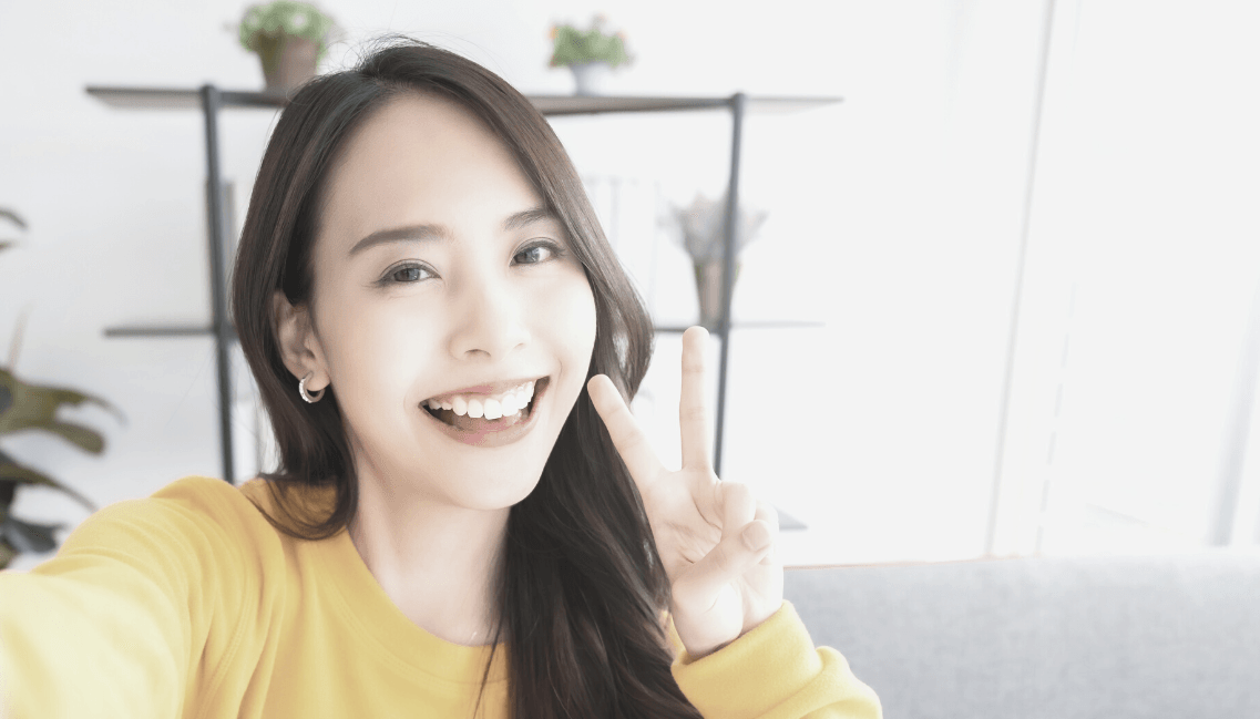 A woman in a yellow sweater is taking a selfie and giving a peace sign.