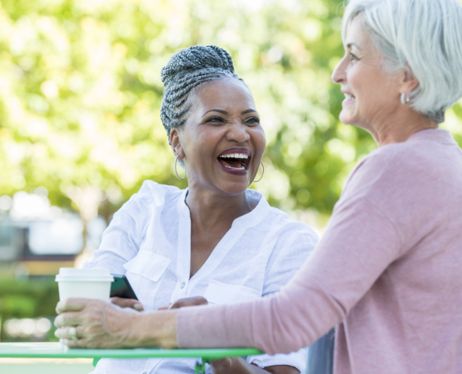 Two older women are sitting at a table laughing and drinking coffee.