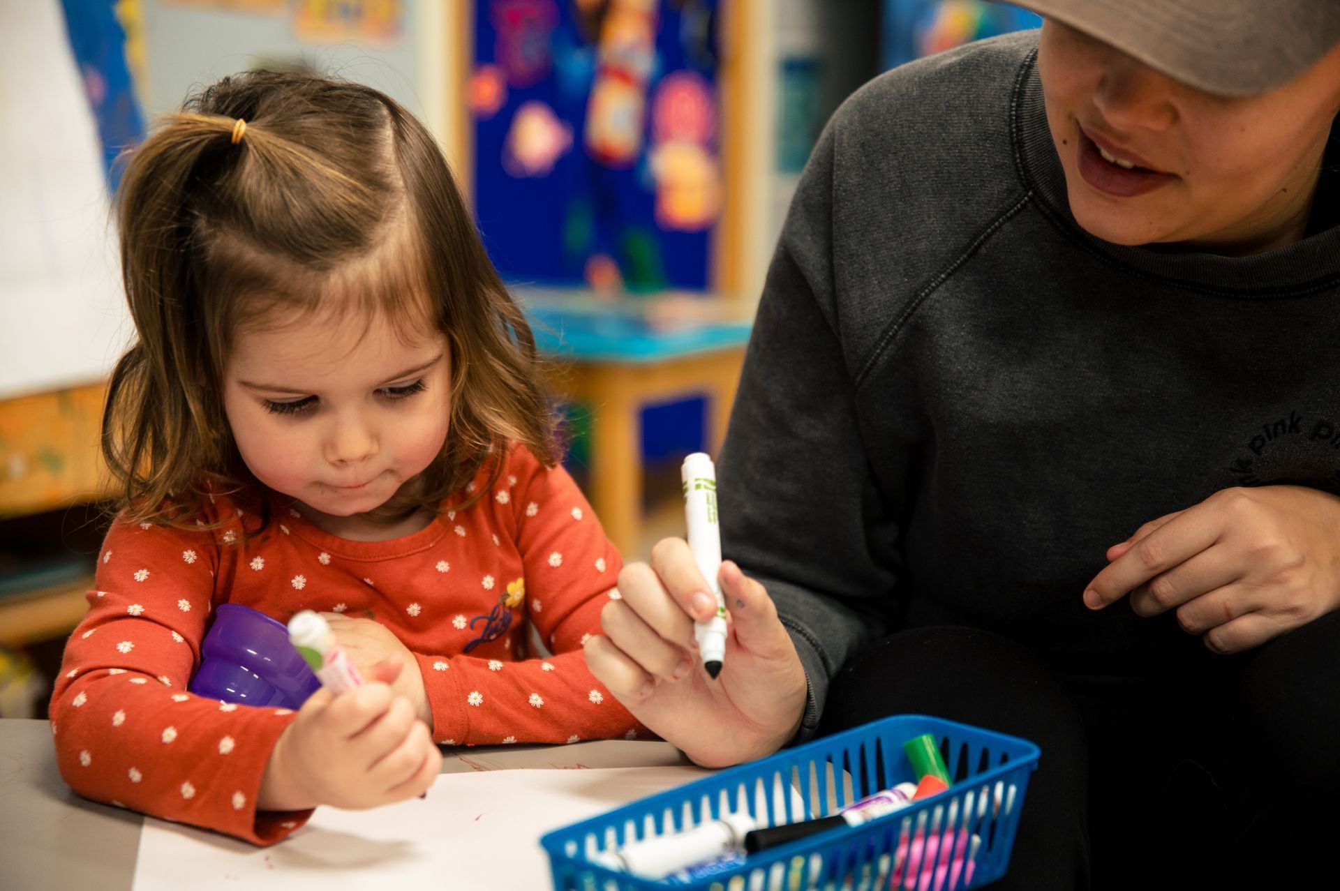 Girl with markers drawing at table with a person; classroom setting.