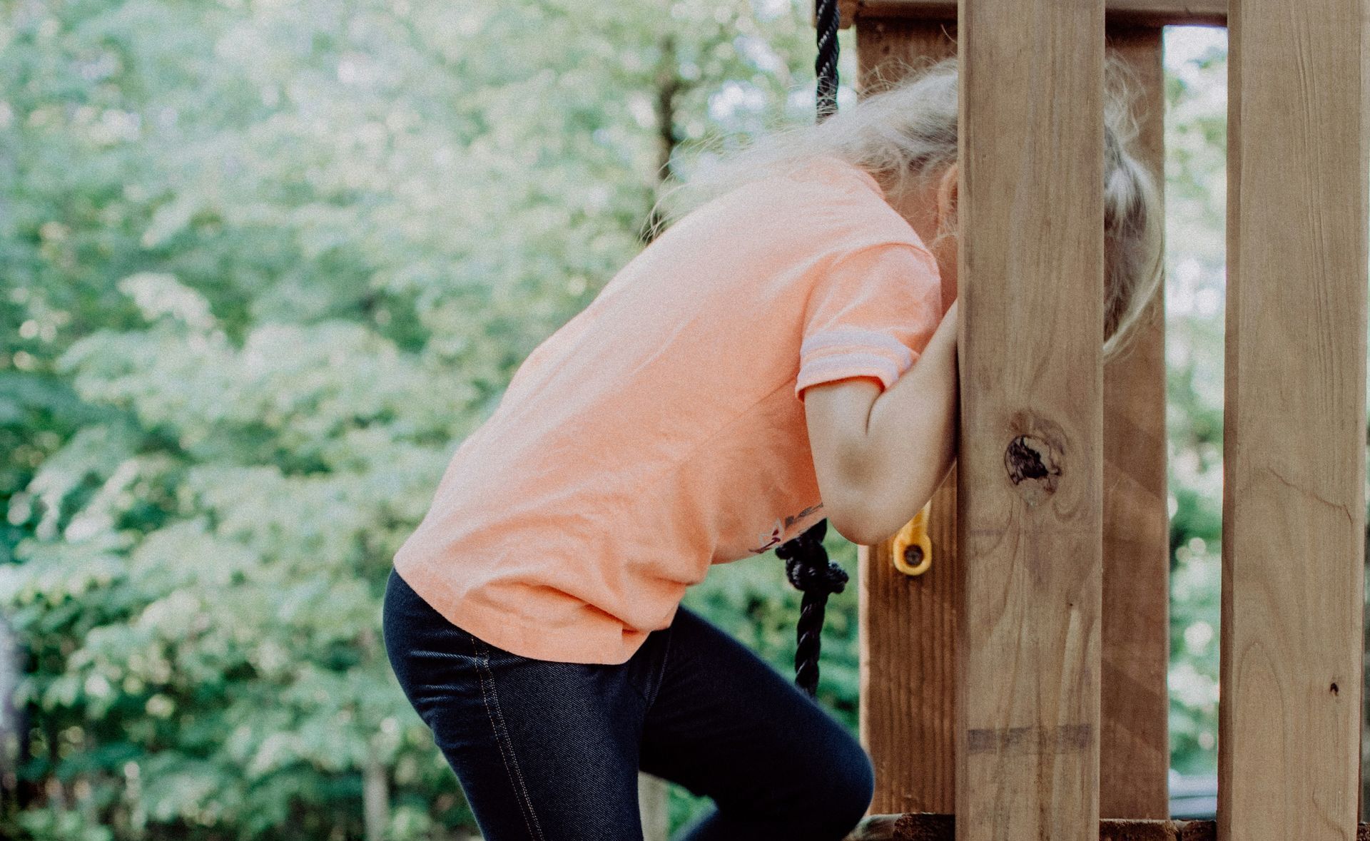 Child peeks out from wooden play structure, wearing orange shirt and jeans.