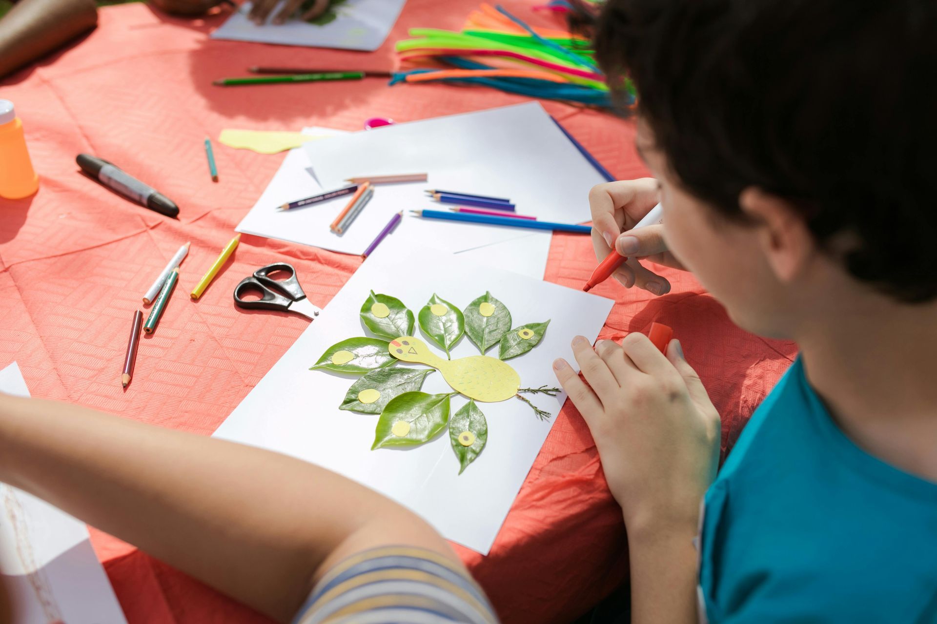 Boy drawing on artwork with leaves, scissors, crayons, and glue on table.