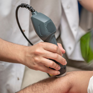 A practitioner uses a handheld acoustic therapy device on a patient's forearm in a clinical office setting.