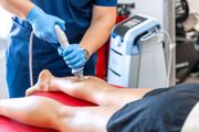 A healthcare professional in blue scrubs uses a handheld shockwave therapy device on a patient's lower leg.
