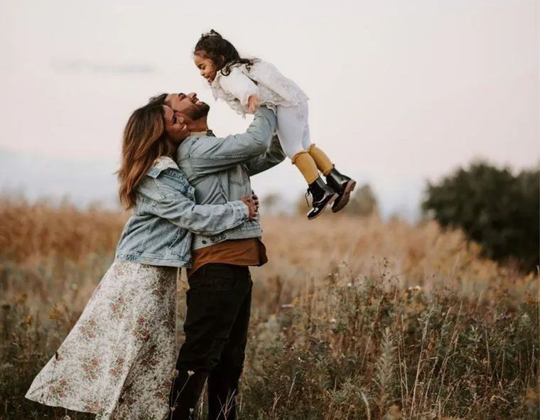 A parent lifts a young child into the air while another adult embraces them in a golden field at sunset.