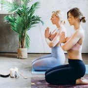 Two people kneeling on mats in a room with a plant and candles, hands held together in a prayer pose, eyes closed.