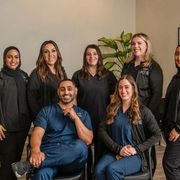 Seven staff members in professional scrubs and dark tops pose for a portrait in a bright office space with a small plant.