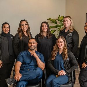 Seven staff members in professional scrubs and dark tops pose for a portrait in a bright office space with a small plant.