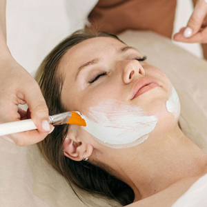 A professional uses a brush to apply a brown skincare mask to a person’s face at a spa.