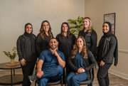 Seven staff members in professional scrubs and dark tops pose for a portrait in a bright office space with a small plant.