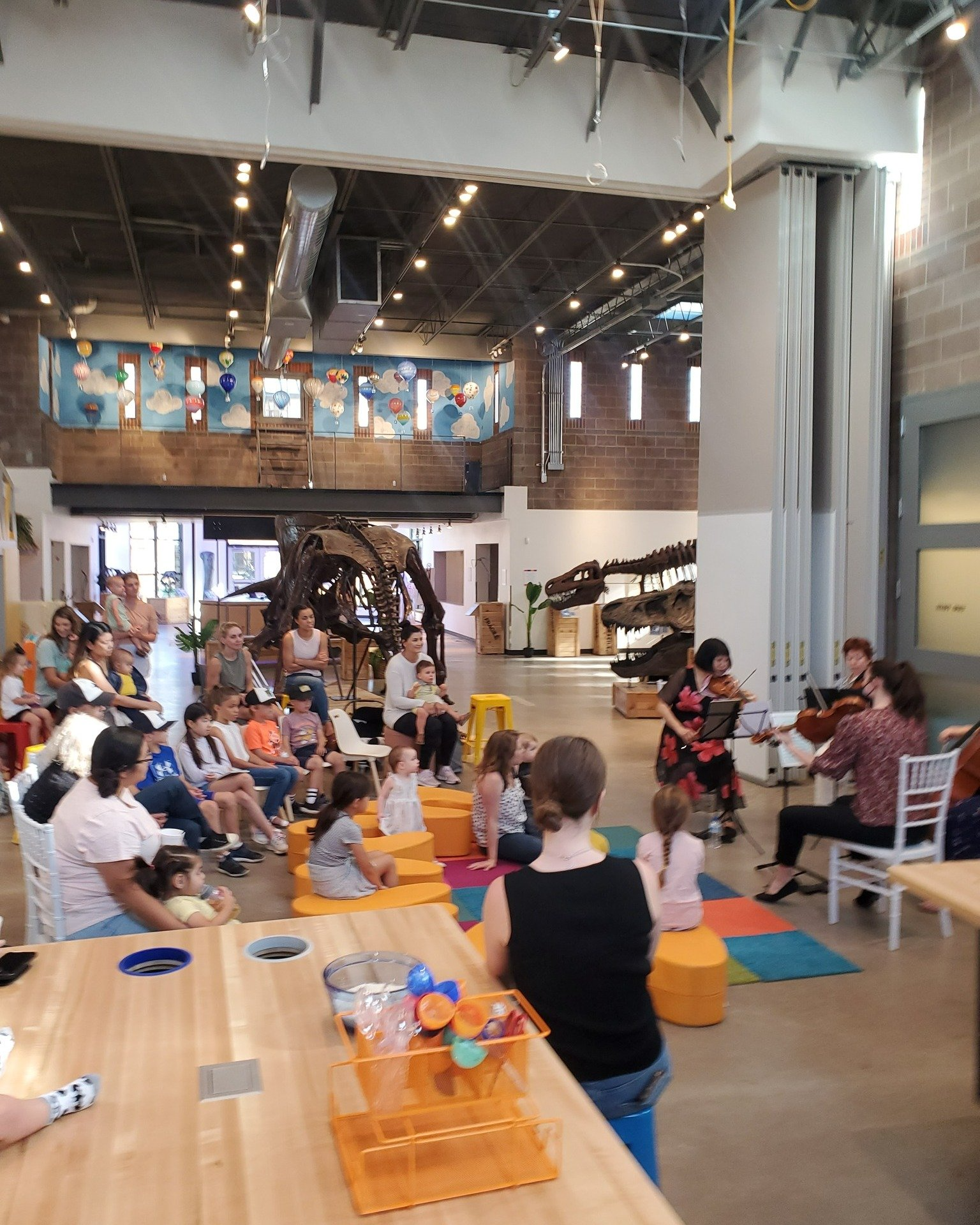 People seated watching musicians playing in a museum. A dinosaur skeleton is in the background.