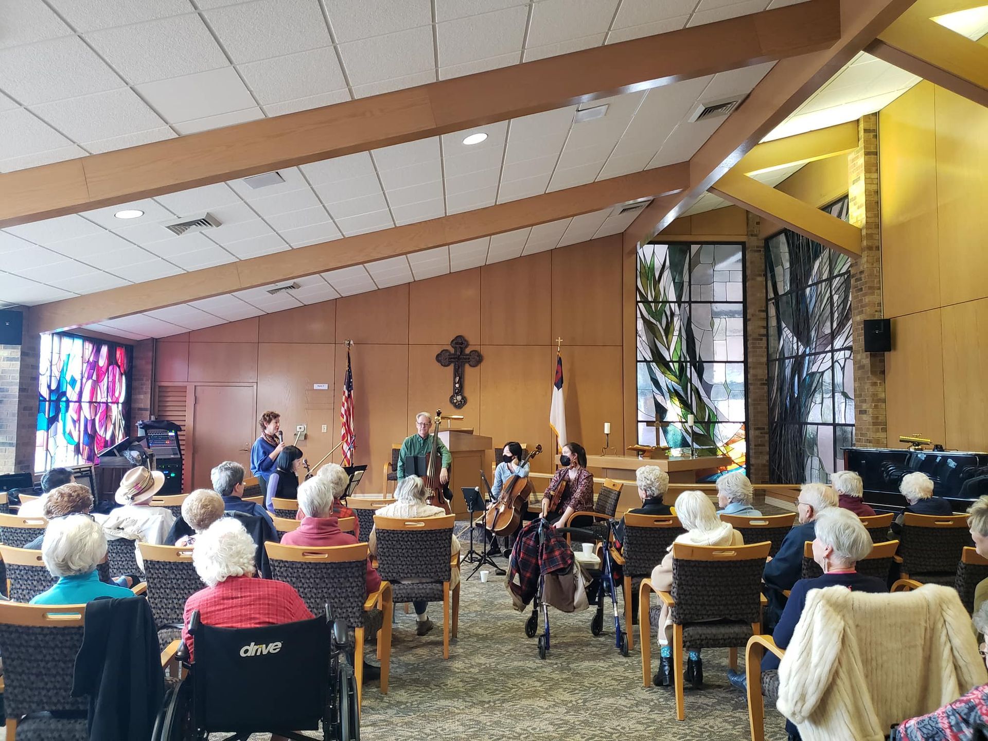 Audience seated, listening to musicians in a room with stained glass and cross; wood paneling on walls.