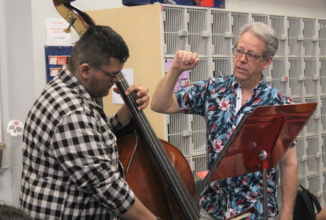 Man plays upright bass as instructor gestures with music stand in a room.