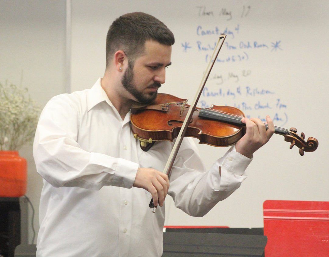 Man playing a violin in a room, white shirt, bow in hand, whiteboard background.