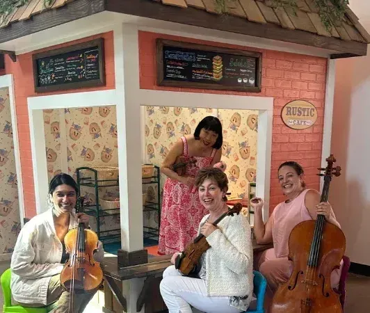 Four musicians with string instruments pose in front of a toy cafe.