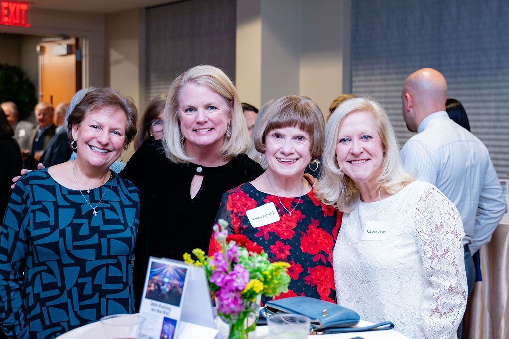 Four women at an event smiling, posing at a table with flowers.