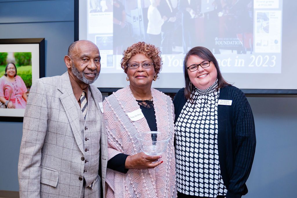 Three people smiling in a room; a man in a tan suit, a woman in a shawl, and another woman in a black and white top.