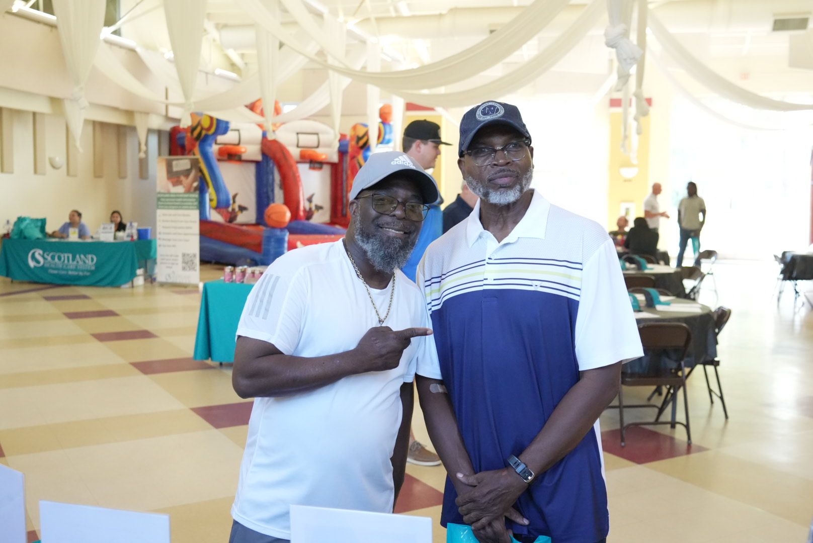 Two men smiling, one pointing. Indoor event with bounce house and tables.